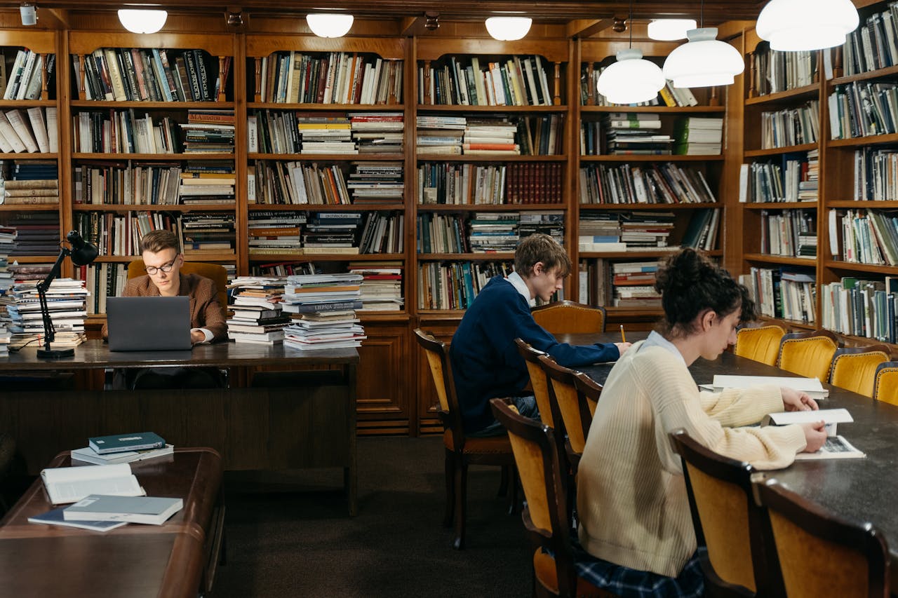 People reading and studying in a cozy library with classic bookshelves and warm lighting.